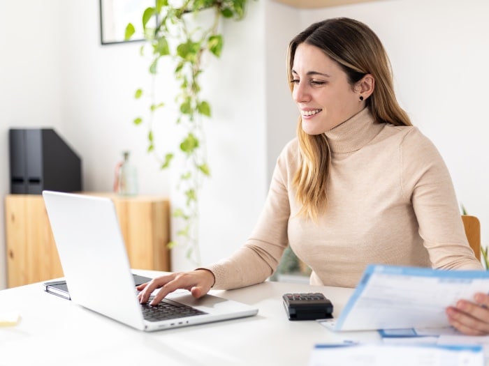 Smiling young woman consulting business invoices sitting on desk at home office workplace